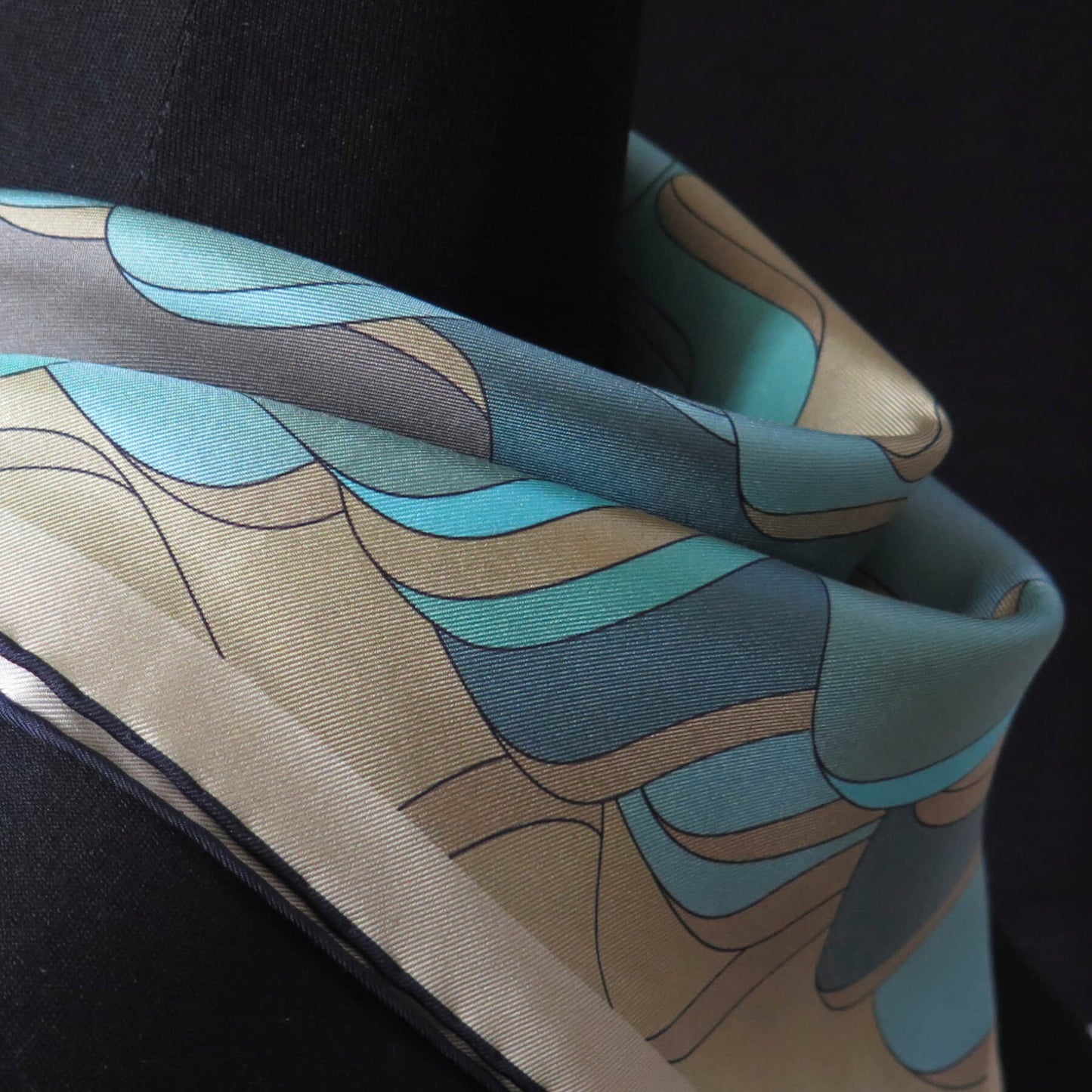 Close up of pure silk scarf with abstract patter in gold, teal, light blue, turquoise and brown, tied in a loose neckerchief style on a tailors dummy