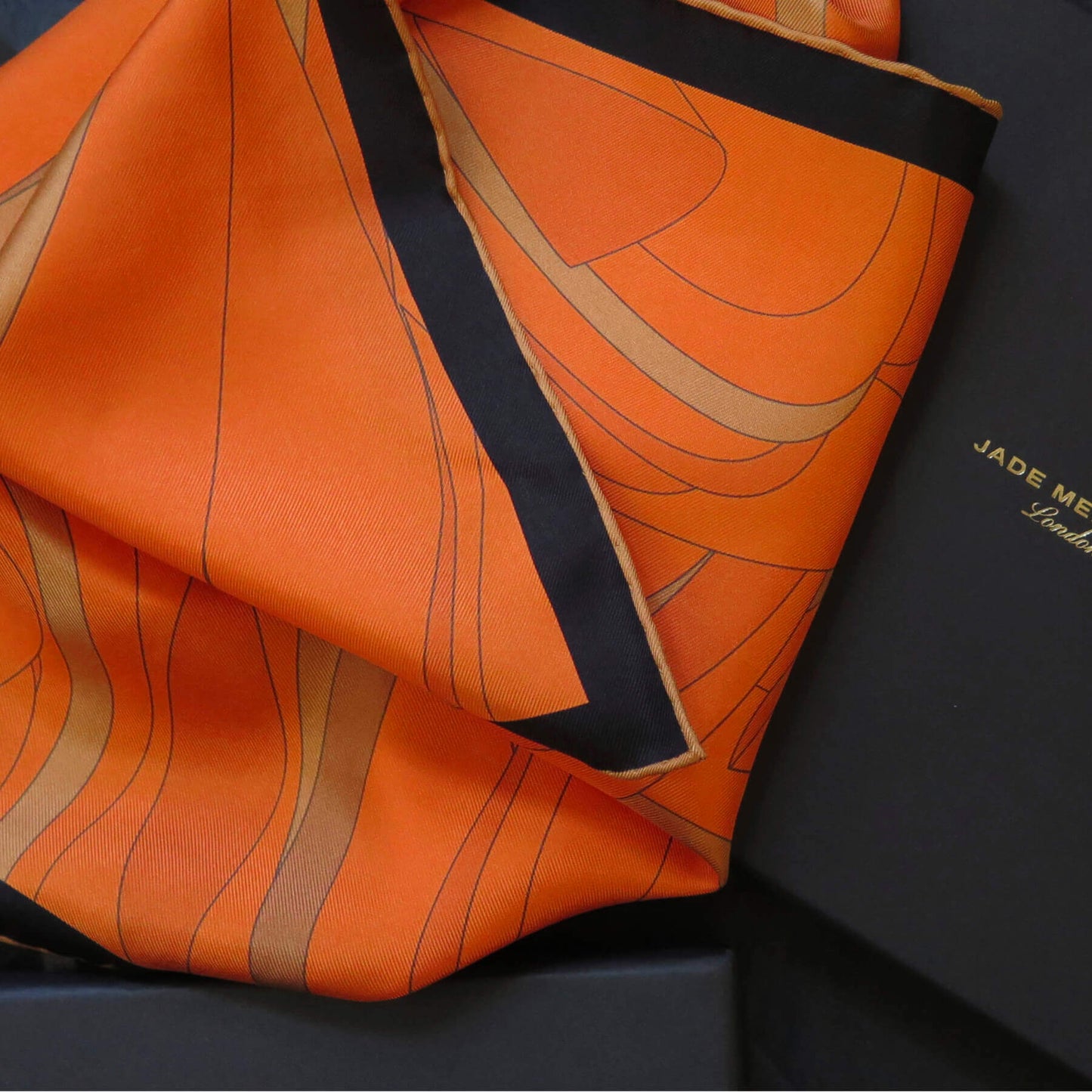 Image of orange and gold pure silk scarf with a dark border and hand-rolled bronze hem. Placed on a branded luxury gift box