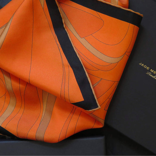 Image of orange and gold pure silk scarf with a dark border and hand-rolled bronze hem. Placed on a branded luxury gift box