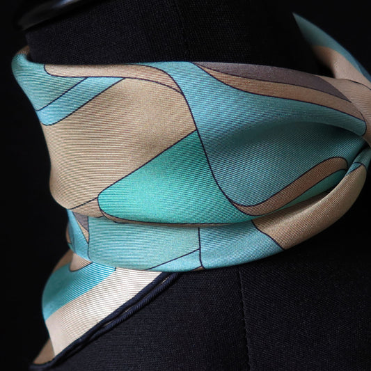 Close up of pure silk scarf with abstract patter in gold, teal, light blue, turquoise and brown, tied in a neckerchief style on a tailors dummy